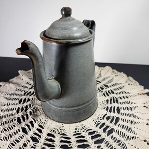 Puede incluir: Una tetera de esmalte gris con asa negra y tapa. La tetera está sobre un mantel de ganchillo blanco.
