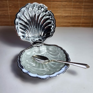 Puede incluir: Una caja de baratijas plateada en forma de concha con una tapa con bisagras, abierta para revelar un interior de color claro. Una pequeña cuchara plateada descansa dentro de la caja. La caja tiene un borde decorativo con cuentas y un acabado pulido.