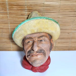 May include: A wall-mounted sculpture of a person's head wearing a wide-brimmed yellow sombrero with a green trim. The figure has a brown complexion, a dark mustache, and a cigar in the mouth, and is wearing a red neckerchief around the neck.
