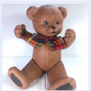 May include: A brown teddy bear ornament with a tartan bow tie. The bear has jointed limbs and black detailing on its paws. It is posed with its arms raised, giving a friendly appearance.