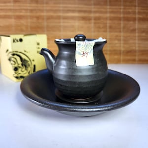 Peut inclure: Une théière en céramique noire avec un petit bec et un couvercle, posée sur une soucoupe assortie. Une petite étiquette est attachée à la théière. Une boîte jaune avec un graphique est en arrière-plan.