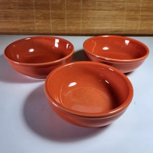 Op de afbeelding: Drie oranje-rode keramische kommen. De kommen zijn rond met een licht uitlopende rand. De binnenkant van de kommen is glad en glanzend en reflecteert licht. De kommen zijn in een groep gerangschikt, met één op de voorgrond en twee op de achtergrond.