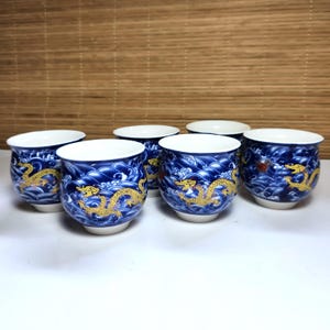 May include: Six blue and white ceramic tea cups with a dragon design. The cups have a white interior and a blue exterior with a wave pattern and a gold dragon. The cups are arranged in a semi-circle.