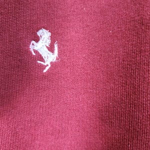 May include: Close-up of a maroon fabric with a white embroidered prancing horse logo. The logo is positioned in the upper left corner of the image. The fabric has a ribbed texture.