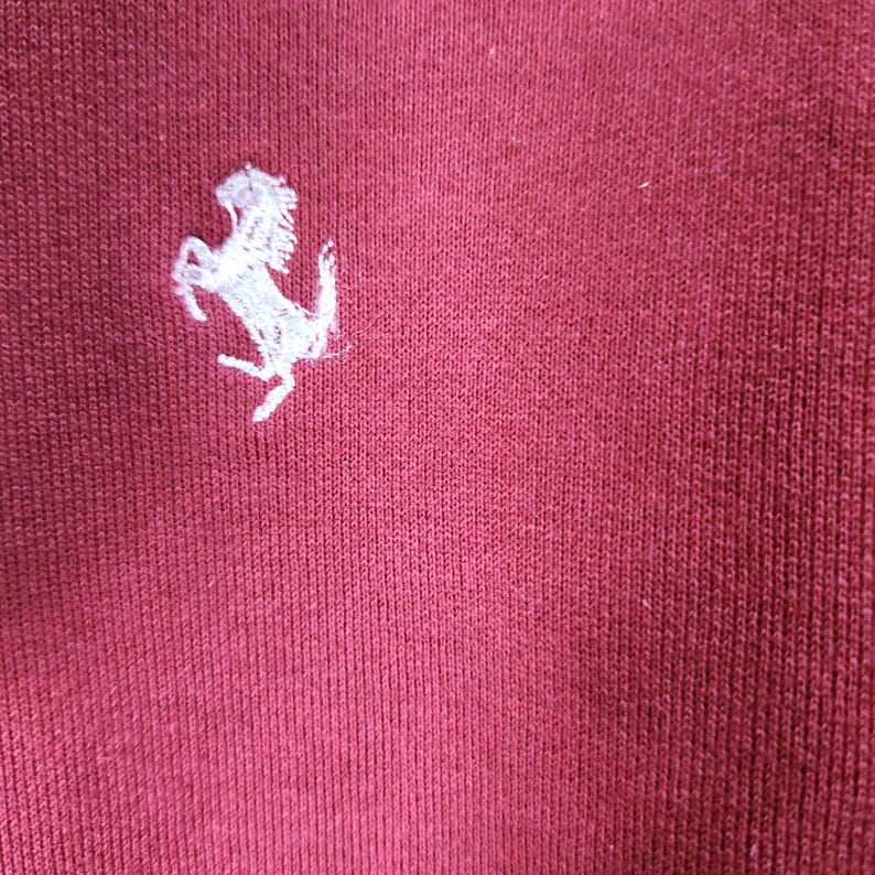 May include: Close-up of a maroon fabric with a white embroidered prancing horse logo. The logo is positioned in the upper left corner of the image. The fabric has a ribbed texture.
