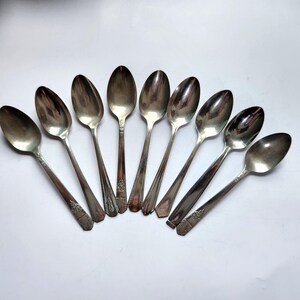 Könnte beinhalten: Eine Sammlung von acht Vintage-Silberlöffeln, die auf einer weißen Oberfläche angeordnet sind. Die Löffel haben ovale Schalen und aufwendige Verzierungen an den Griffen. Die Löffel bestehen aus einem glänzenden, reflektierenden Metall.