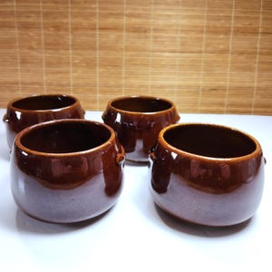 May include: A set of four small, round, dark brown ceramic bowls with a glossy finish. Each bowl features a wide opening, a smooth curved body, and two small, protruding handles on opposite sides, arranged in a group on a white surface.