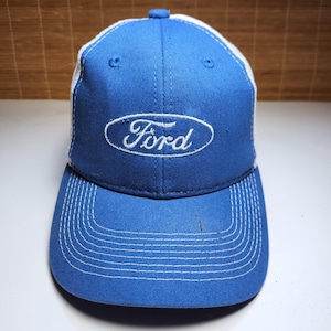May include: A blue baseball cap with a white mesh back and a white embroidered Ford logo in an oval on the front. The cap features a curved brim with white stitching detail and a button on the top center, providing a classic structured look.