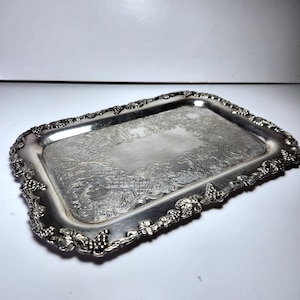 May include: A rectangular silver tray with detailed floral and grape vine edging. The tray has a reflective surface, likely metal. The design is vintage and elegant, with a decorative, ornate pattern.