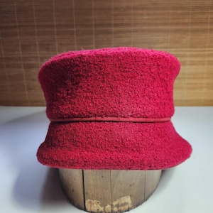 May include: A vibrant red cloche hat with a textured, wool-like appearance. The hat features a rounded crown and a slightly flared brim, with a thin band detail. The hat is displayed on a wooden form.