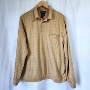 May include: Tan long-sleeve shirt with a collared neckline and a button-down placket. The shirt features a grid pattern and a chest pocket. The shirt is hanging on a wooden hanger.