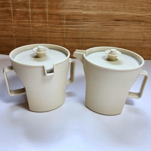 Può includere: Due contenitori in plastica color bianco sporco con coperchi e manici abbinati. Un contenitore presenta un beccuccio laterale per versare, mentre l'altro ha un bordo dentellato. Entrambi i recipienti hanno una finitura opaca e liscia.