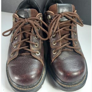 May include: A pair of dark brown leather lace-up boots with a rounded toe, featuring metal eyelets, brown laces, and a black padded collar. The tongue of the boot displays the CAT logo in black and white text. The boots have a thick black sole.