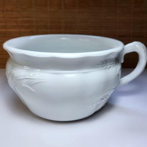 May include: A white ceramic chamber pot featuring a wide, rounded bowl with a smooth, glossy finish and a single curved handle on the side. The exterior surface is decorated with a subtle, embossed wheat stalk pattern encircling the body.