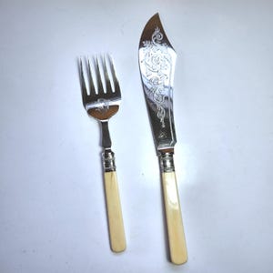 Puede incluir: Un tenedor y un cuchillo de pescado plateados con mangos color crema. La hoja del cuchillo tiene un diseño floral ornamentado. El tenedor tiene cuatro púas. Los mangos son de color crema claro.