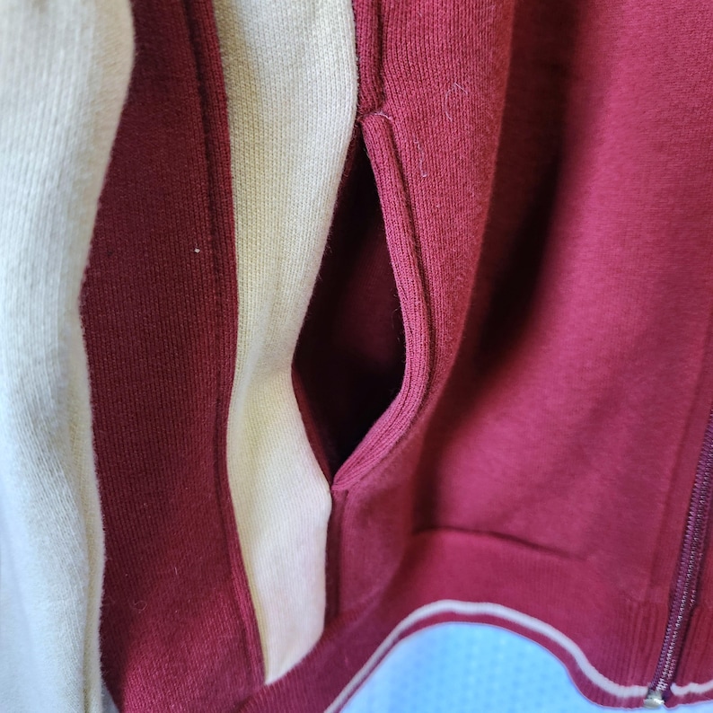 May include: Close-up of a maroon zip-up jacket with cream-colored stripes down the side. The jacket has a pocket and a white trim along the bottom. The zipper is silver.