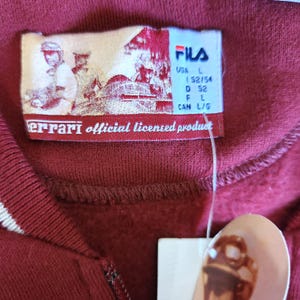 May include: Close-up of a burgundy Fila sweatshirt with a white stripe detail. The tag reads "Ferrari official licensed product" and includes the Fila logo and size information. The sweatshirt is a size L.