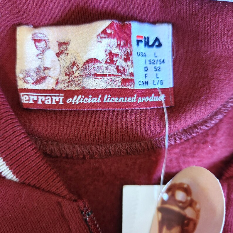 May include: Close-up of a burgundy Fila sweatshirt with a white stripe detail. The tag reads "Ferrari official licensed product" and includes the Fila logo and size information. The sweatshirt is a size L.