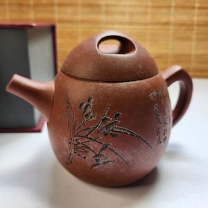 May include: A round, matte brown clay teapot featuring a textured surface with an engraved black botanical illustration of grass or bamboo on the front and vertical black Chinese calligraphy characters on the side, with a circular handle lid.