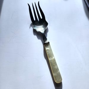 May include: A silver serving fork with four tines and a decorative handle. The handle is a light cream colour with a pearlescent sheen. The fork is designed for serving and has a classic, elegant appearance.