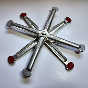 Puede incluir: Una herramienta de neumáticos Zephyr plateada vintage con seis brazos que irradian desde un cubo central. Cada brazo tiene una almohadilla roja y circular en el extremo. La herramienta está marcada con la palabra "ZEPHYR" y "MARQUE DEPOSEE".