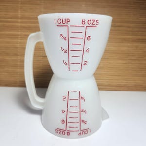 May include: White glass measuring jug with a handle. The jug has red measurement markings for cups and fluid ounces. The text "1 CUP" and "8 OZS" are printed on the jug. The jug is designed to measure up to 237 ml.