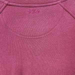 May include: Close-up of a maroon sweatshirt with the brand name "FILA" embroidered in a matching color. The sweatshirt has a crew neck and raglan sleeves. The fabric appears to be a soft knit.