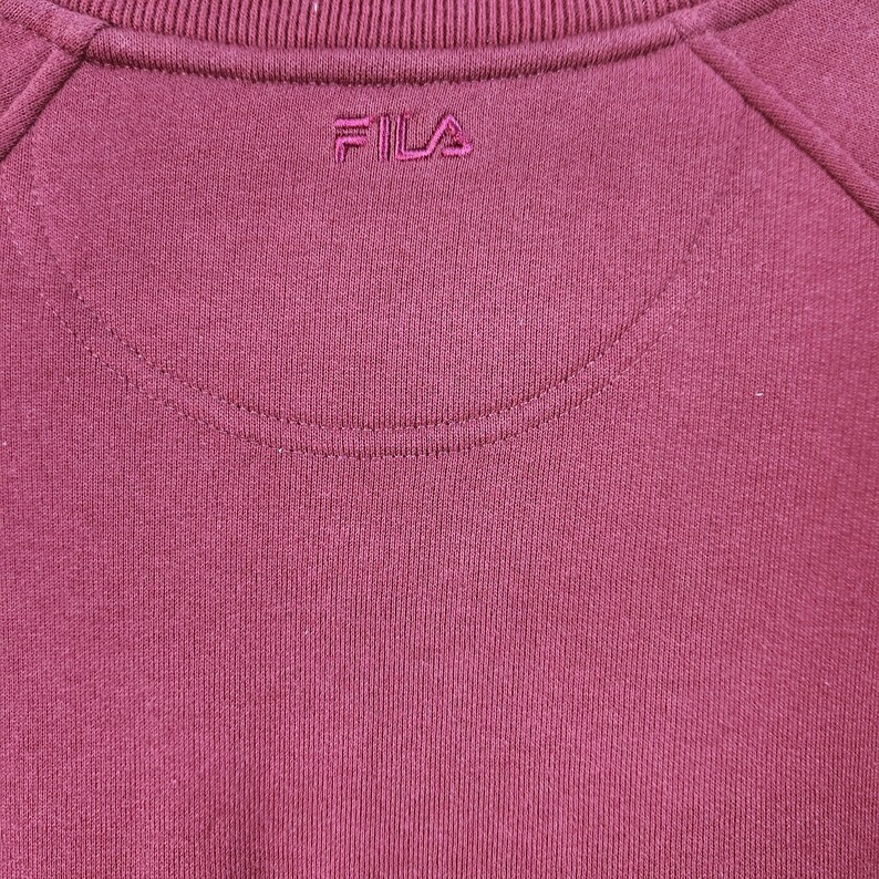 May include: Close-up of a maroon sweatshirt with the brand name "FILA" embroidered in a matching color. The sweatshirt has a crew neck and raglan sleeves. The fabric appears to be a soft knit.