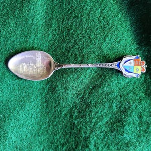 Puede incluir: Cuchara de té de recuerdo de plata con una representación del edificio del Parlamento canadiense en el cuenco y un escudo de esmalte colorido con el escudo de armas canadiense en el mango. La cuchara está sobre un fondo verde.