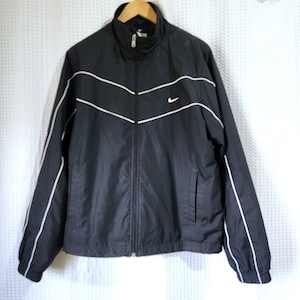 Puede incluir: Una chaqueta deportiva negra con cremallera, cuello alto, mangas largas con puños elásticos y detalles de ribetes blancos en el pecho y a lo largo de los brazos. Un pequeño logotipo Nike swoosh blanco está bordado en la parte superior izquierda.