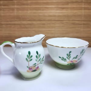 May include: A white ceramic creamer jug and sugar bowl set. The jug has a green handle and a floral design with green leaves and pink flowers. The sugar bowl has a matching design and a gold rim. The set is on a white surface.
