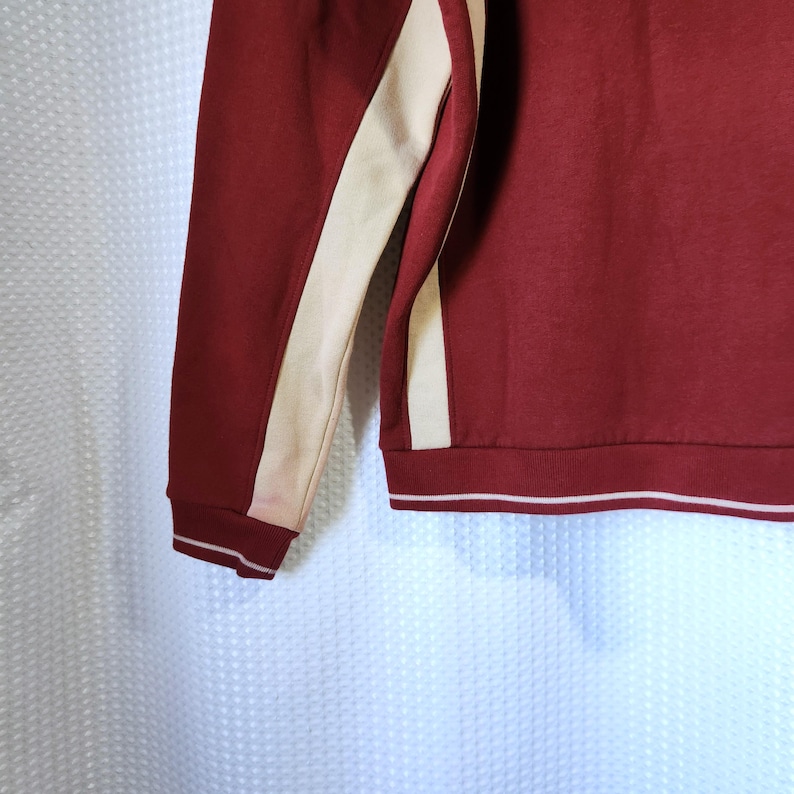 May include: Maroon sweatshirt with cream-colored stripes down the sleeves. The cuffs and hem have a white stripe detail. The fabric appears to be a soft, knit material. The overall style is casual and sporty.