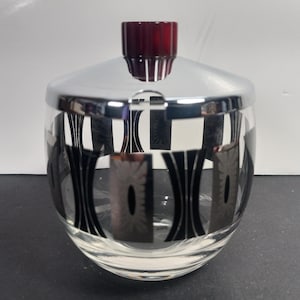 Puede incluir: Un tarro de cristal transparente con tapa plateada y tapón rojo oscuro. El tarro tiene un patrón geométrico negro. El tarro está sobre una superficie negra.