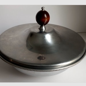 Könnte beinhalten: Eine silberfarbene Servierschüssel mit Deckel und einem dunkelbraunen Holzknauf. Die Schüssel hat einen Perlenrand und eine abgerundete Form. Die Metalloberfläche reflektiert das Licht.