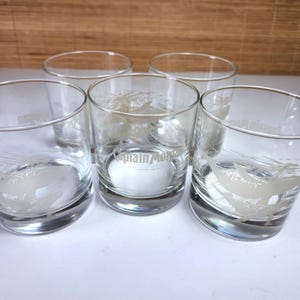 以下が含まれることがあります： 丸底の透明なグラス5個セット。各グラスには鳥の彫刻と「Captain Morgan」の文字が施されています。グラスは白い表面に配置されています。