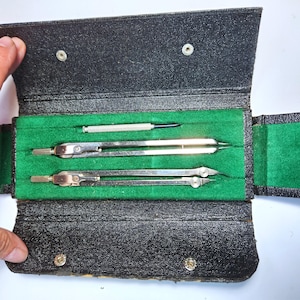 May include: A vintage drafting set in a black leatherette case with a green velvet lining. The set includes two silver-coloured compasses and a small screwdriver. The case is open, revealing the tools inside.