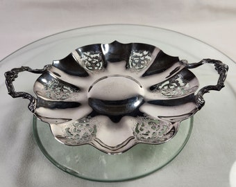 May include: A silver-colored, scalloped dish with ornate handles. The dish has a circular center and decorative floral patterns around the rim. The handles have a detailed, scroll-like design. The dish is sitting on a clear glass surface.