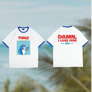 Op de afbeelding: Witte T-shirts met blauwe bies en een ronde hals. Eén shirt heeft een cartoon tonijn met het woord "TUNA" erboven. Het andere shirt heeft de tekst "DAMN, I LOVE FISH" en "FISHING (1992) SOCIETY".
