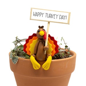 May include: A terracotta-colored flower pot with a decorative turkey figurine. The turkey has a brown body, a red wattle, and a colorful fan of feathers. It holds a sign that reads "HAPPY TURKEY DAY!" in black letters.