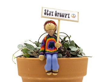 May include: A small clay figurine of a hippie wearing a tie-dye shirt and blue jeans sits on the edge of a terracotta pot. The figurine is holding a sign that says "Stay Groovy" with a peace sign.