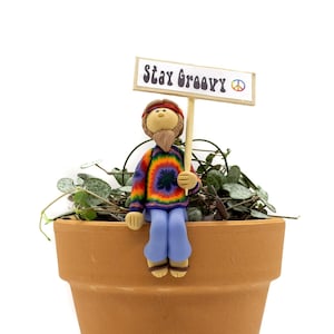 May include: A small clay figurine of a hippie wearing a tie-dye shirt and blue jeans sits on the edge of a terracotta pot. The figurine is holding a sign that says "Stay Groovy" with a peace sign.