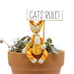May include: A clay figurine of an orange and white tabby cat holding a sign that says "CATS RULE!" The cat is sitting on the edge of a terracotta pot with a green plant growing inside.