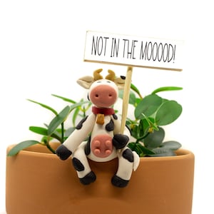 May include: A white and black cow figurine holding a sign that says "NOT IN THE MOOOOD!" The cow is sitting on the edge of a terracotta pot with a green plant growing inside.
