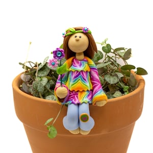 May include: A handcrafted polymer clay figurine of a girl in a colorful rainbow-patterned top, blue pants, and a flower crown. She holds a flower and sits in a terracotta pot with a green plant.