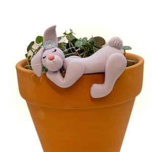 May include: A light purple bunny figurine with a blue flower on its head, resting on the rim of a terracotta pot filled with green plants. The bunny has a pink nose and a small, fluffy tail. The pot is a classic, unglazed design.