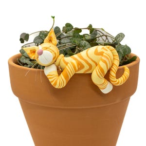 May include: A terracotta-colored flower pot with a String of Hearts plant. A whimsical, orange and yellow striped cat figurine is draped over the pot's edge, appearing to nap. The cat has white paws and a pink nose.
