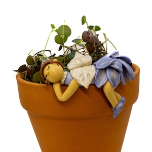May include: A terracotta-colored flower pot with green plants and a whimsical fairy figurine. The fairy is resting on the pot's edge, with a yellow body, brown hair, and a blue and white floral skirt. The fairy is sleeping.