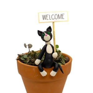 May include: A black and white cat figurine with green eyes and a pink nose, sitting on the rim of a terracotta pot. The cat holds a thin wooden stick topped with a rectangular white sign that features the black text WELCOME in capital letters.