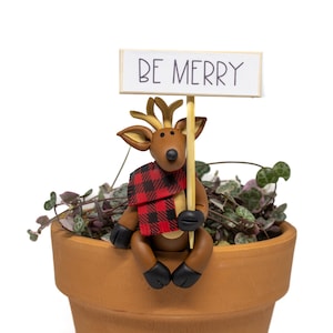 May include: A brown reindeer figurine with a red and black plaid scarf holds a sign that reads "BE MERRY". The reindeer sits on the edge of a terracotta pot filled with a trailing plant. The figurine has black hooves and antlers.