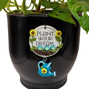 May include: A round white ceramic plant marker with the text "PLANT YOUR DREAM" in black lettering. It features sculpted yellow sunflowers, blue flowers, and green leaves. A blue watering can charm with a sunflower hangs from the bottom.